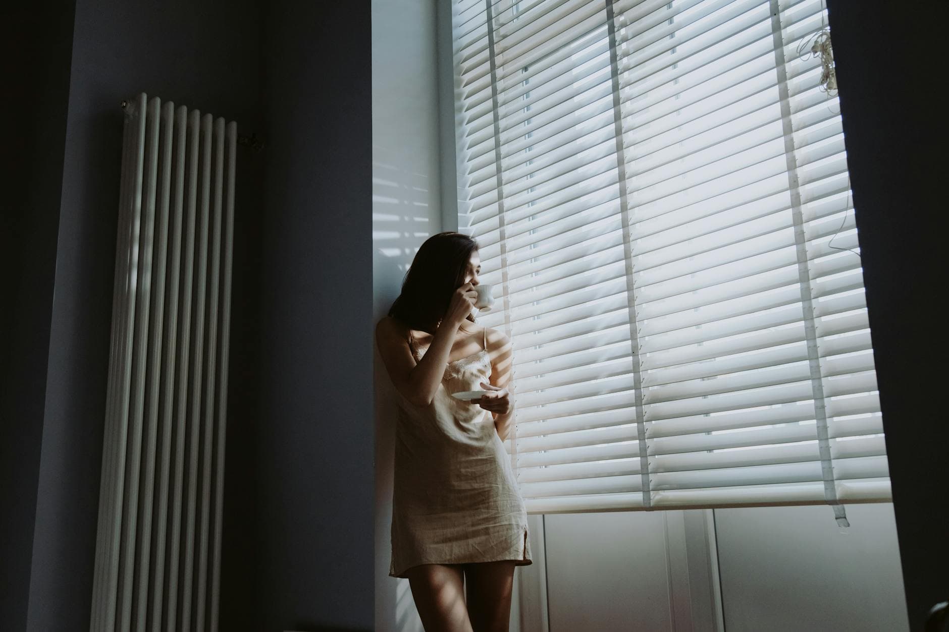 A woman stands by a bright bedroom window in soft morning light, holding a cup of coffee in a quiet moment before starting her day.