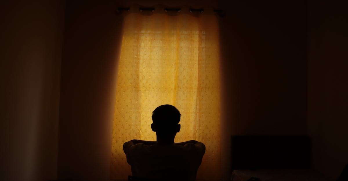 A person sits alone near a window in soft afternoon light, looking out with a distant, unfocused expression.