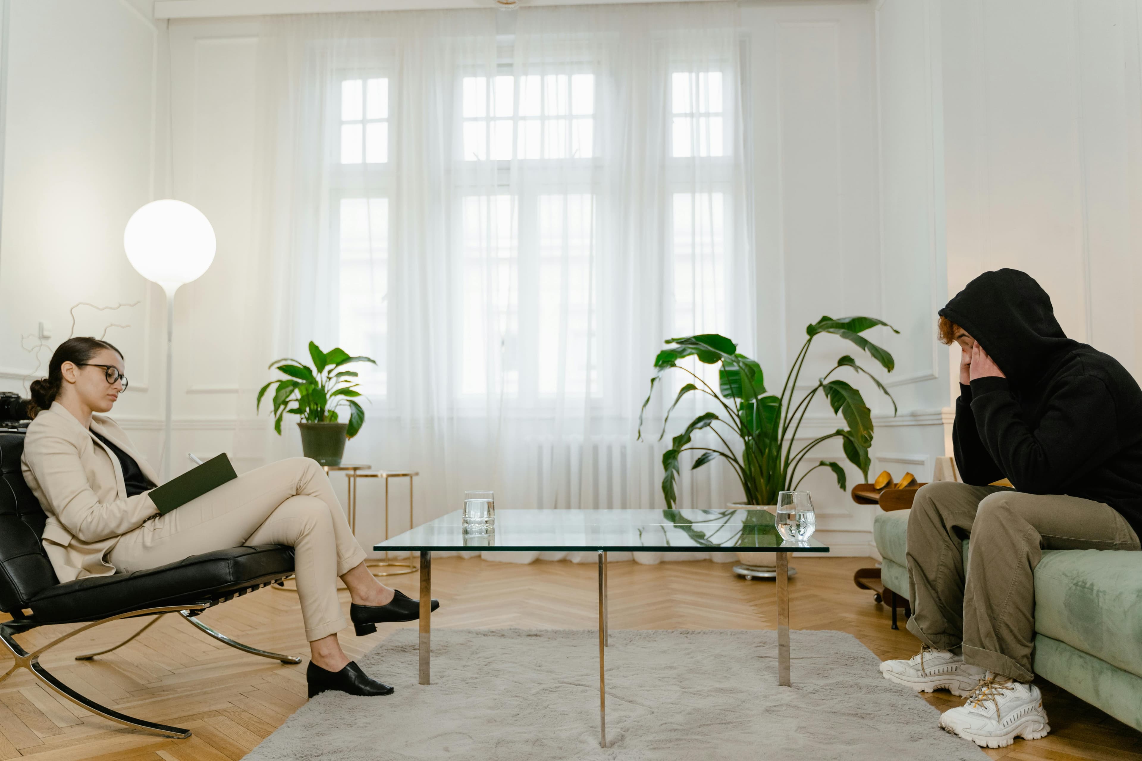 A therapist takes notes while a client sits on a sofa in a bright, calm office during a therapy session.