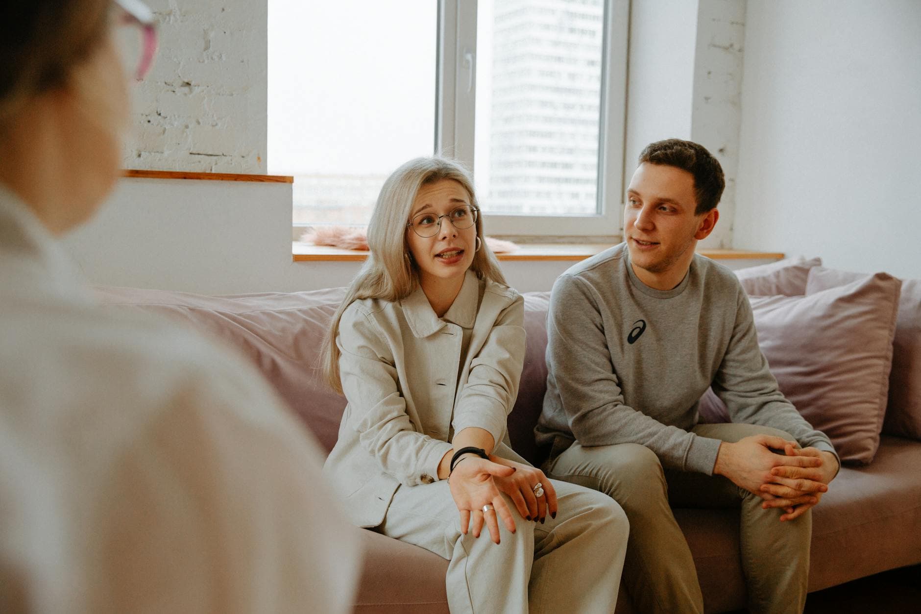 Two partners sit in separate rooms of their California home, each on a laptop, attending an online couples therapy session via video call, with soft natural light coming through the windows.
