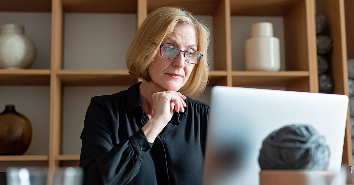 A focused woman in glasses concentrates on her laptop screen at a home desk, appearing absorbed in her work.