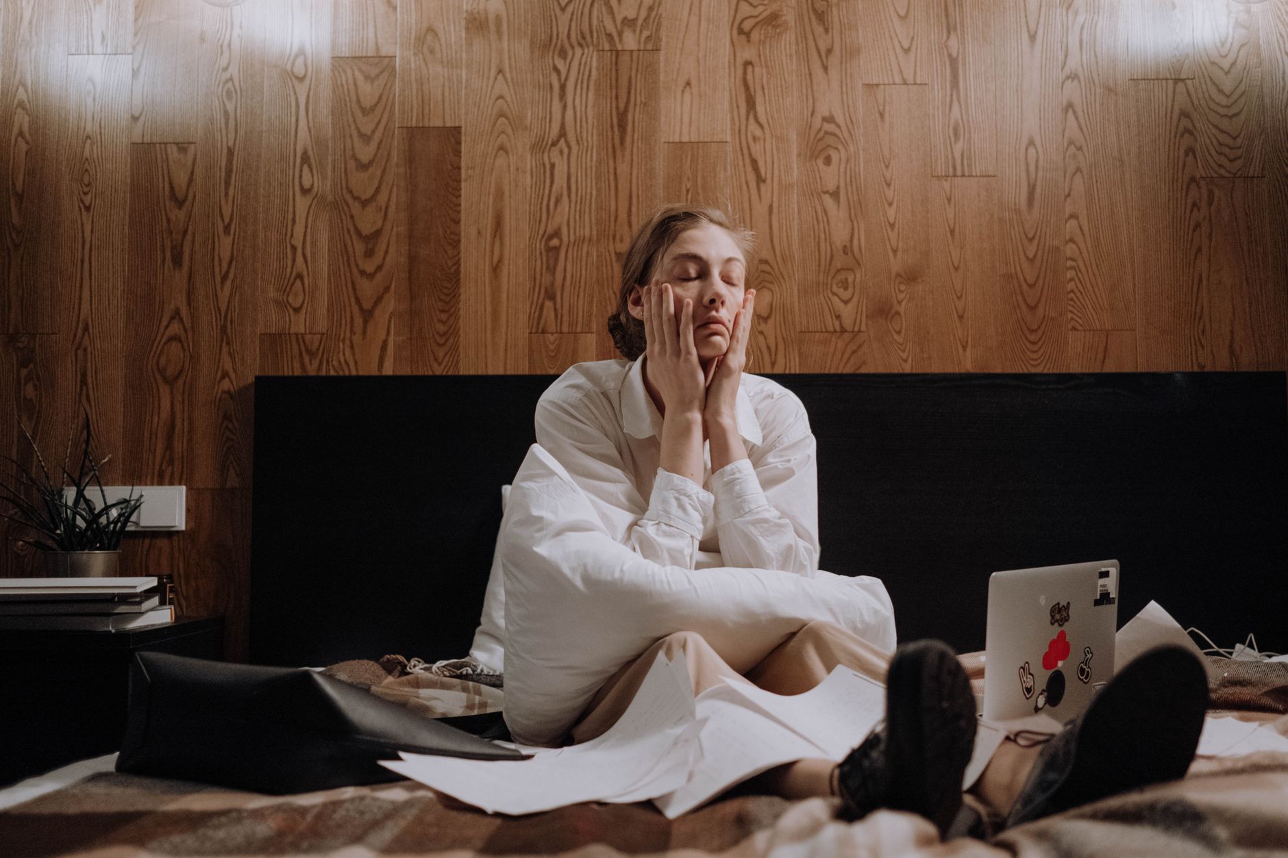 An overwhelmed woman sits on a bed with her hands on her face, unable to focus on the laptop and scattered papers in front of her.