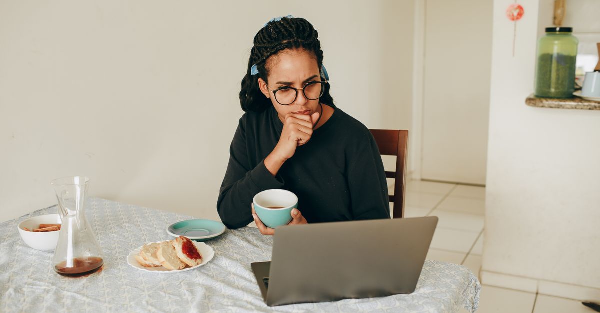 A woman with braided hair and glasses sits at a home dining table holding a mug of coffee, appearing worried and preoccupied as she stares at her laptop next to a plate of bread and jam.