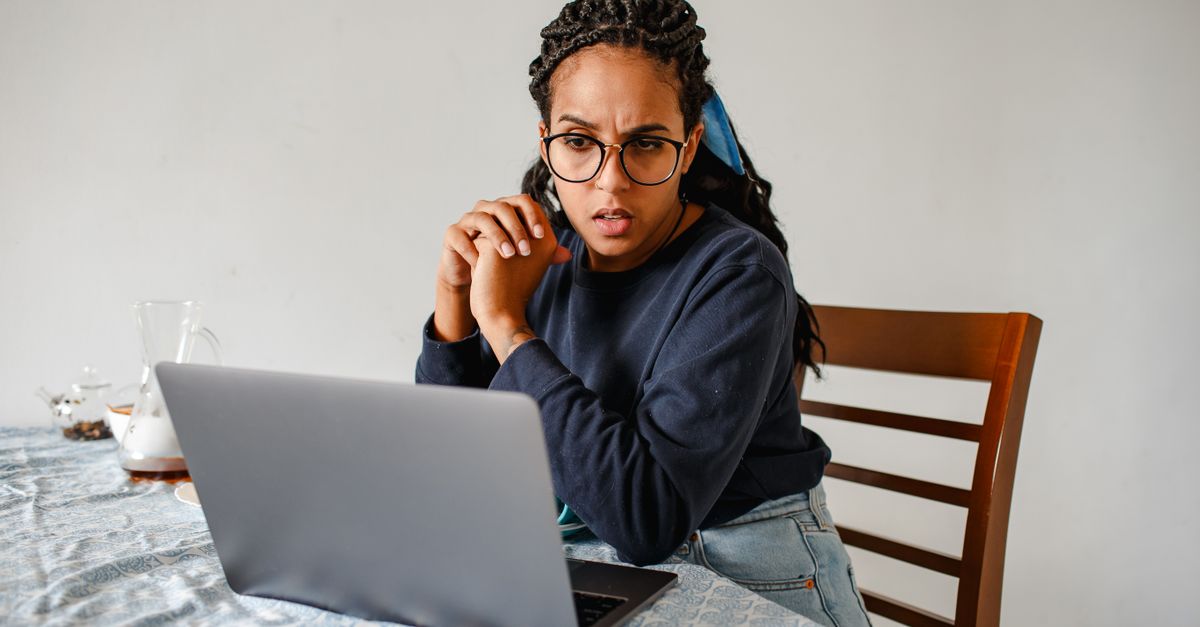 A young woman wearing glasses and a dark sweatshirt sits at a home table, appearing tense and unfocused as she stares at her laptop screen.
