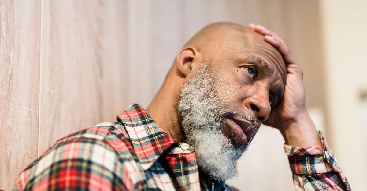 A worried middle-aged man rests his hand on his head with a troubled expression while sitting indoors in a quiet home setting.