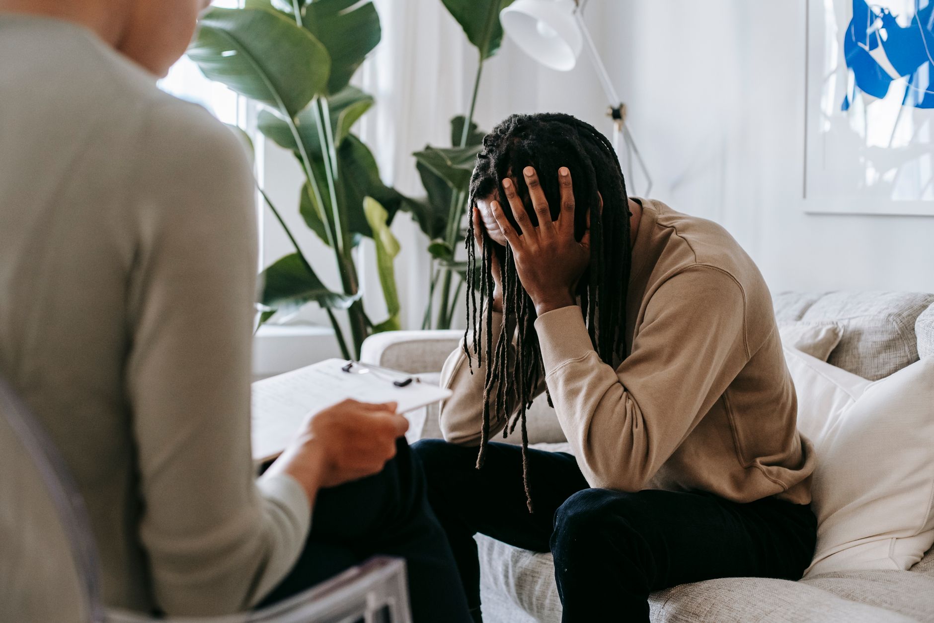 Anxious African American male with hands on head sitting on sofa near crop female psychologist with clipboard during appointment in office.