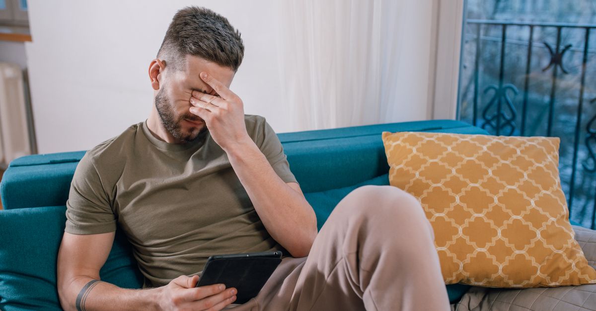 A distressed man sits on a sofa at home, covering his face with his hand while holding a tablet, conveying emotional overwhelm.