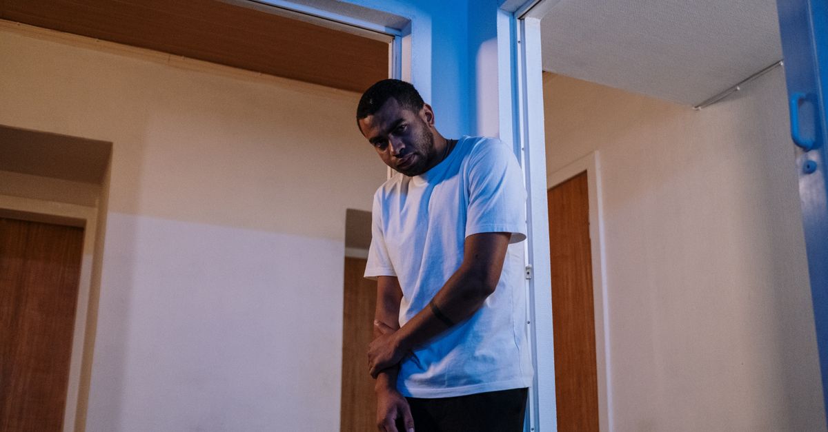 A young man in a white t-shirt stands alone in a dim doorway with his head down, conveying a somber and withdrawn mood.