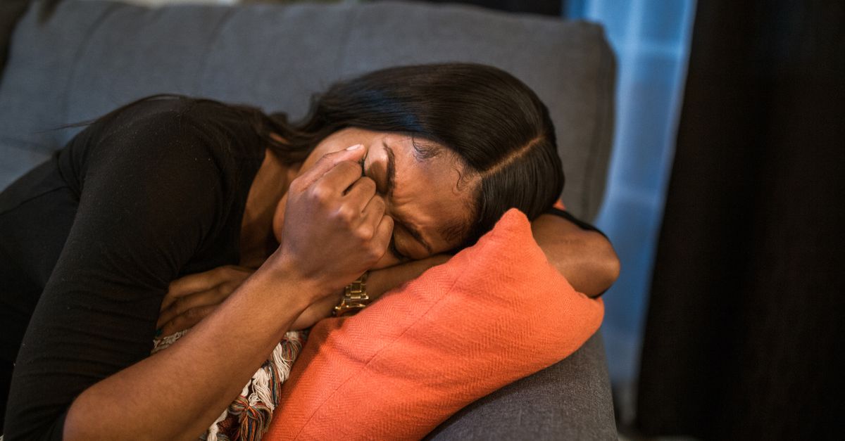 A woman lies curled on a gray couch, clutching an orange pillow and covering her face with her hand as she appears to cry.