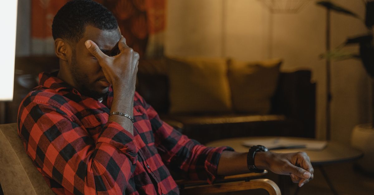 A tired-looking man in a plaid shirt sits slumped in a chair at home, pressing his hand to his forehead in a gesture of fatigue and low mood.