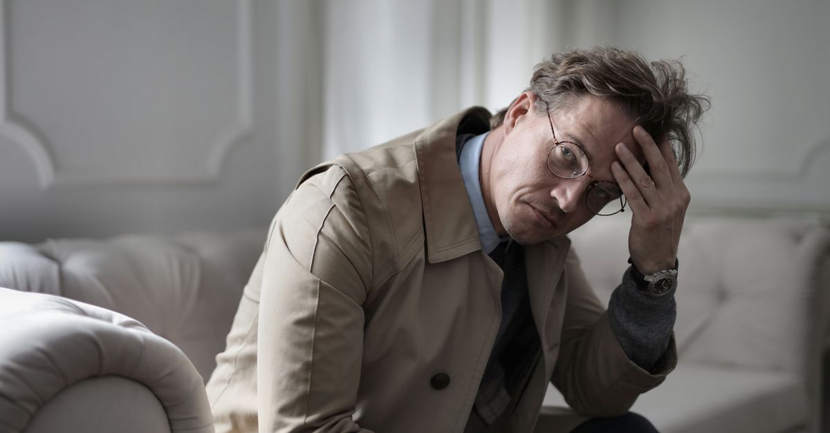 A tired-looking man in glasses and a trench coat sits slumped on a couch at home, holding his head as he stares blankly, conveying low mood and fatigue.