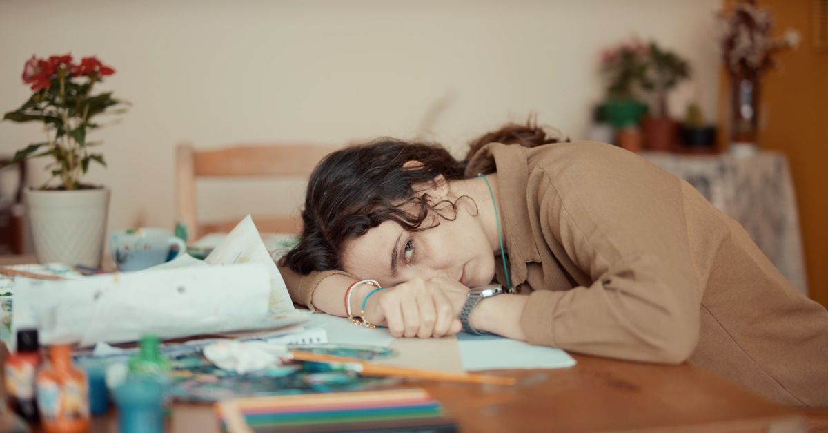 A young woman lies with her head on a desk surrounded by art supplies, staring blankly with a detached, emotionally flat expression.