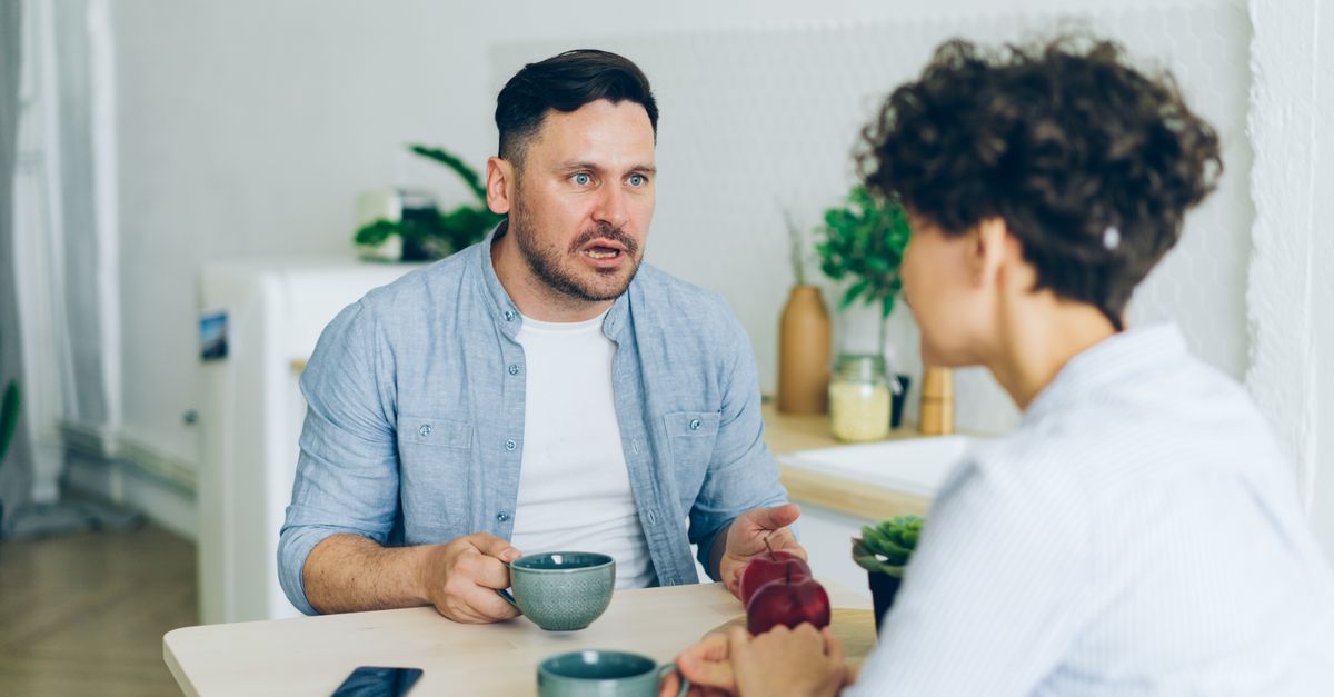A couple having a serious, tense conversation at their kitchen table over cups of coffee.