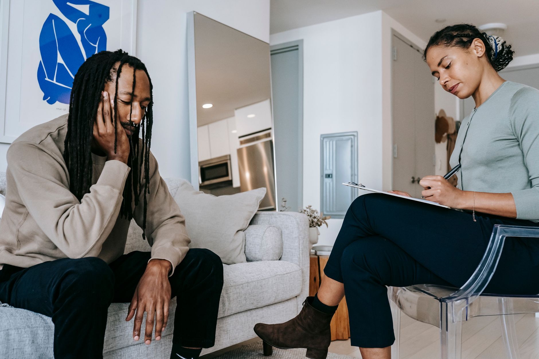 Attentive African American female with clipboard taking notes near sad male partner touching face on couch at home