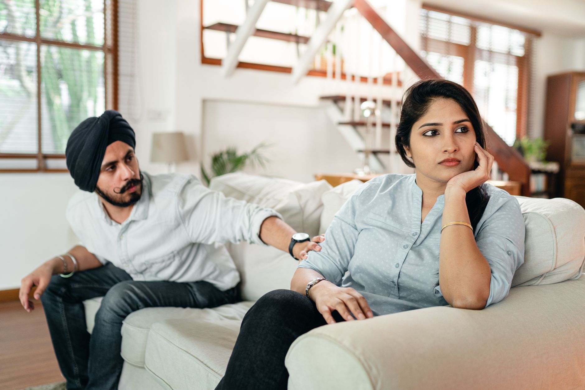 A couple sits on a living room couch having a tense conversation, with the man reaching toward his partner who looks away with a troubled expression.