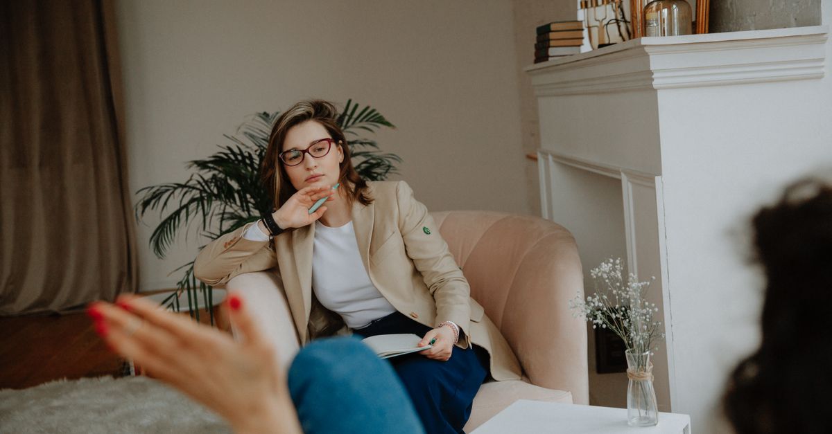 A therapist in glasses and a beige blazer listens attentively while taking notes during a session with a client seated across from her in a cozy room.