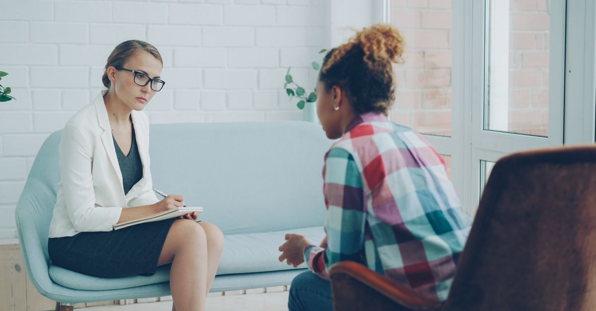 A therapist takes notes while attentively listening to a client during a private counseling session in a bright, calm room.