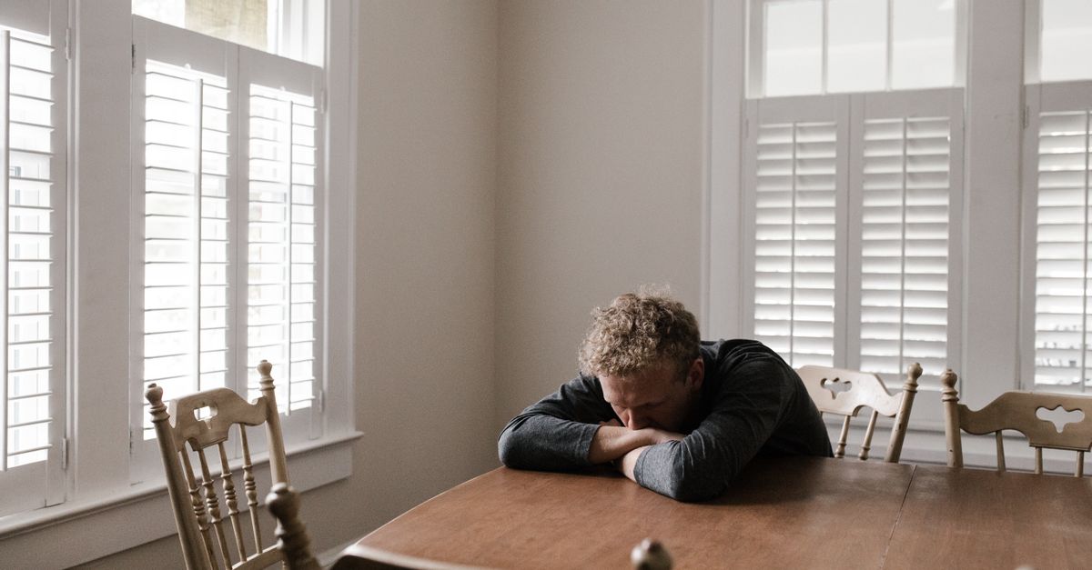 A man sits alone at a wooden table with his head resting on his folded arms, appearing distressed and overwhelmed in a quiet room with shuttered windows.
