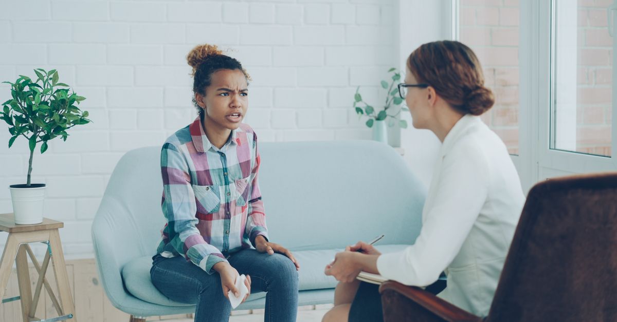 A young woman in a plaid shirt talks with a therapist taking notes during a counseling session in a bright room.