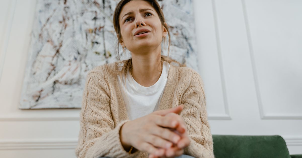 A distressed woman in a cozy beige sweater sits indoors with her hands clasped tightly, showing an anxious and overwhelmed expression.