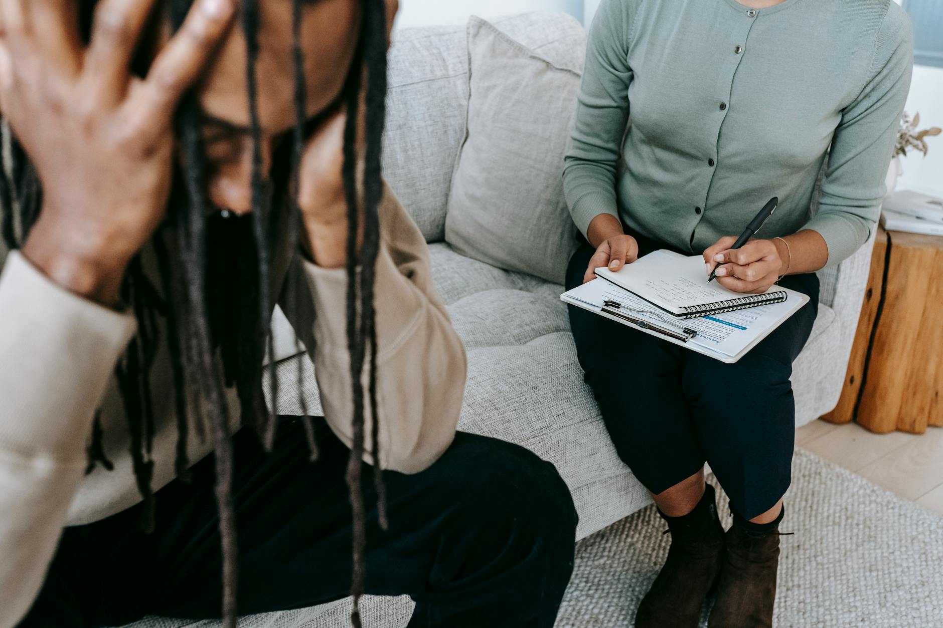 Crop concerned African American male sitting on sofa with female psychologist and holding head in hands in dismay during psychotherapy session.
