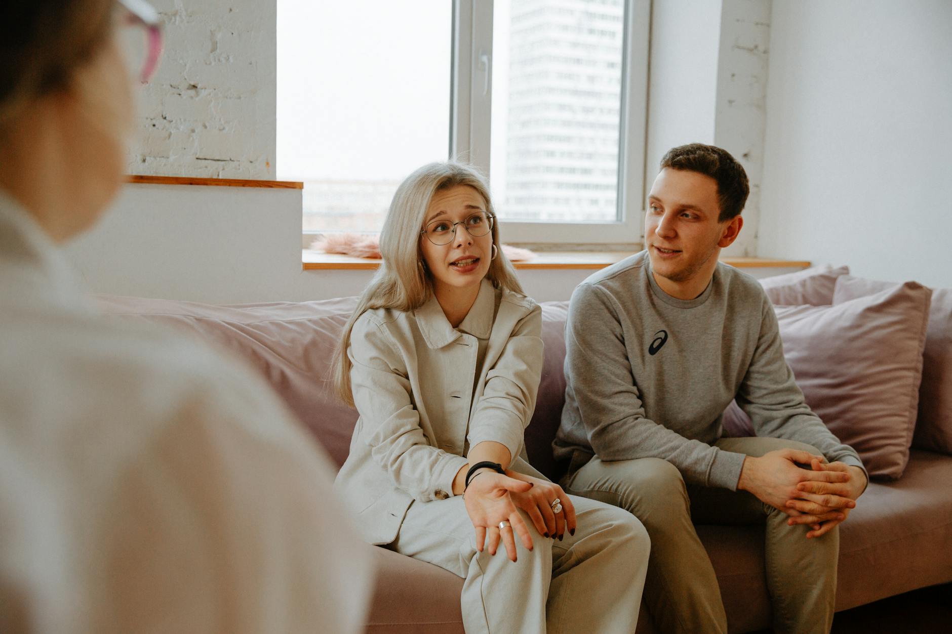 Two partners sit in separate rooms of their California home, each on a laptop, attending an online couples therapy session via video call, with soft natural light coming through the windows.