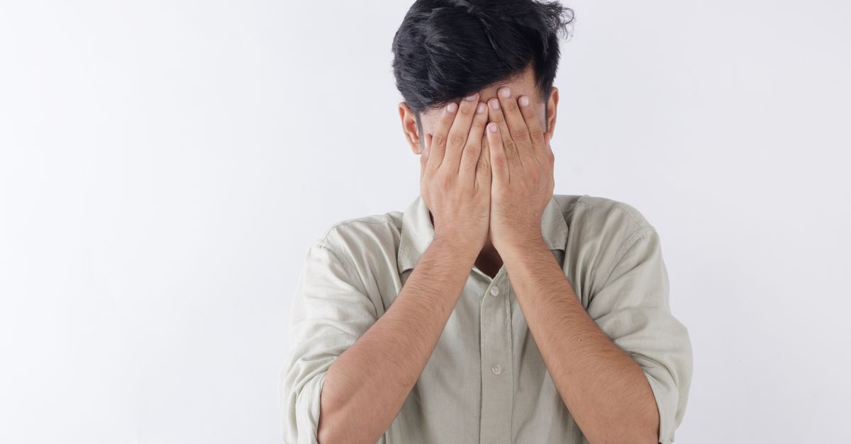 A young man in a light shirt hides his face behind both hands, expressing emotional overwhelm and anxious distress.