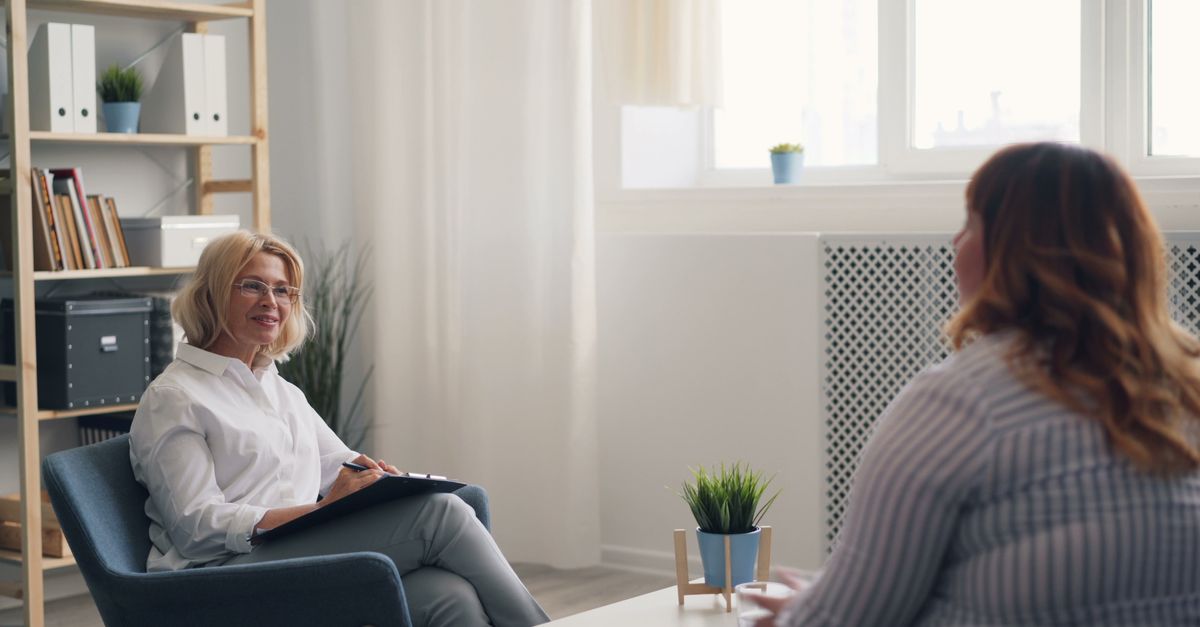 A therapist with a clipboard listens to a client during a counseling session in a bright, modern office.