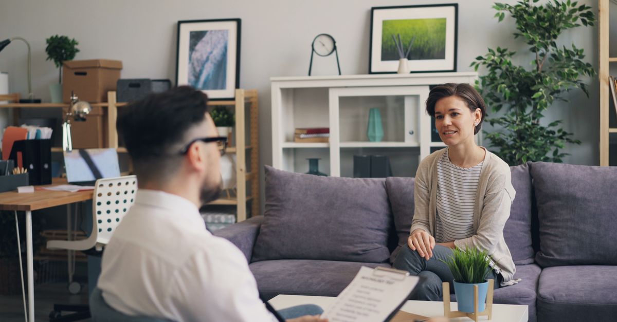 A therapist holding a clipboard conducts an in-person therapy session with a female client seated on a couch in a bright, modern office.