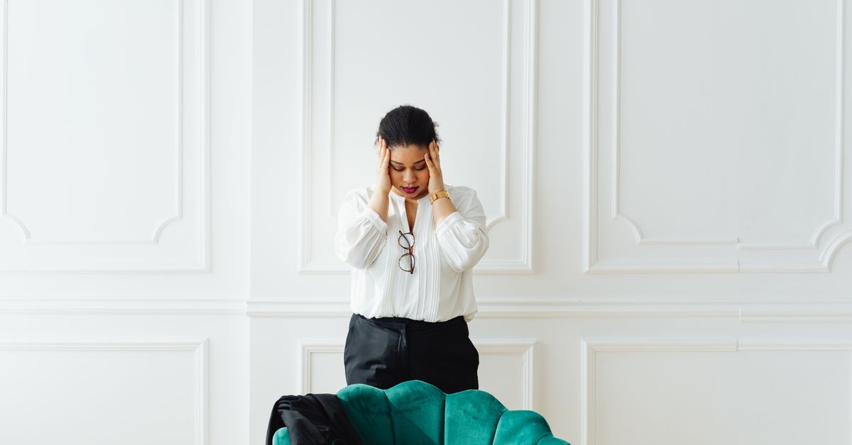 A woman stands in a bright room holding her head in her hands, appearing emotionally overwhelmed and stressed.