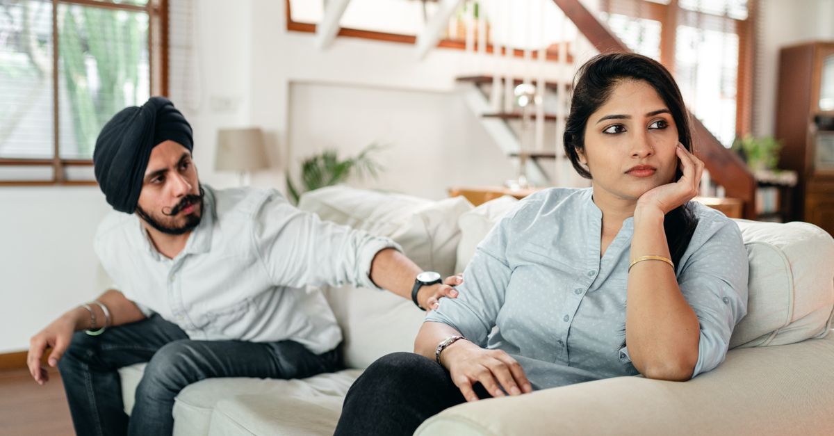 A couple sits on a living room couch during a tense conversation, with the woman looking away while the man reaches toward her.