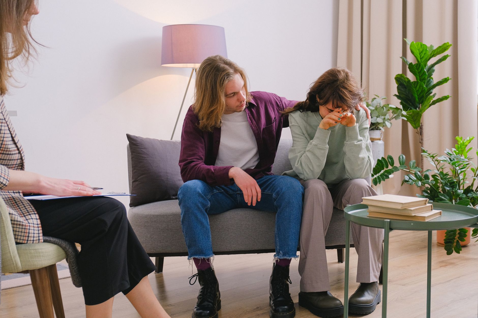 A man embraces his frustrated partner as they speak with a therapist in a bright office.