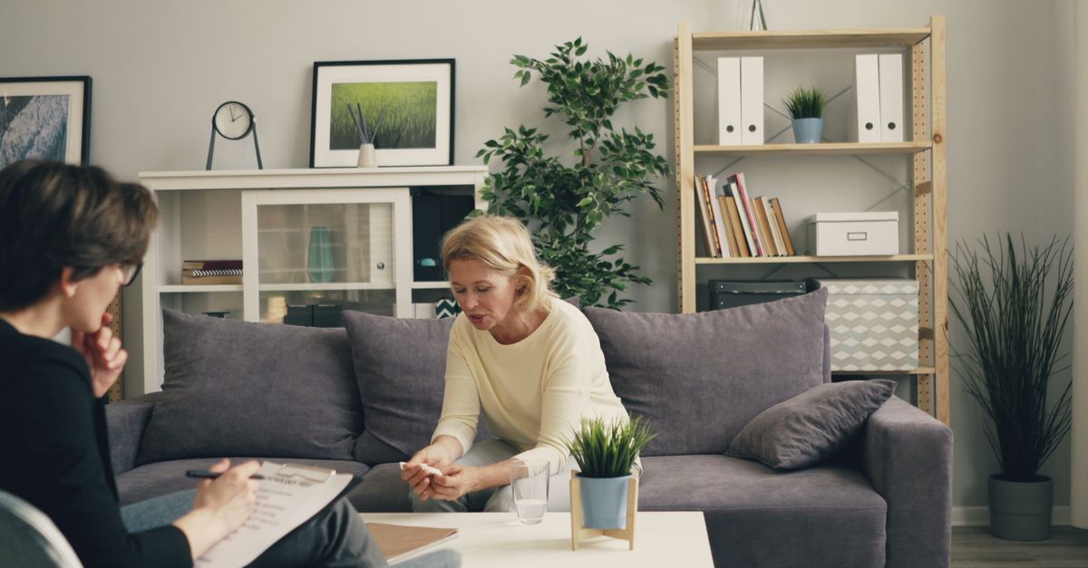 A woman talks with a therapist taking notes on a clipboard during an in-person therapy session in a comfortable living room setting.